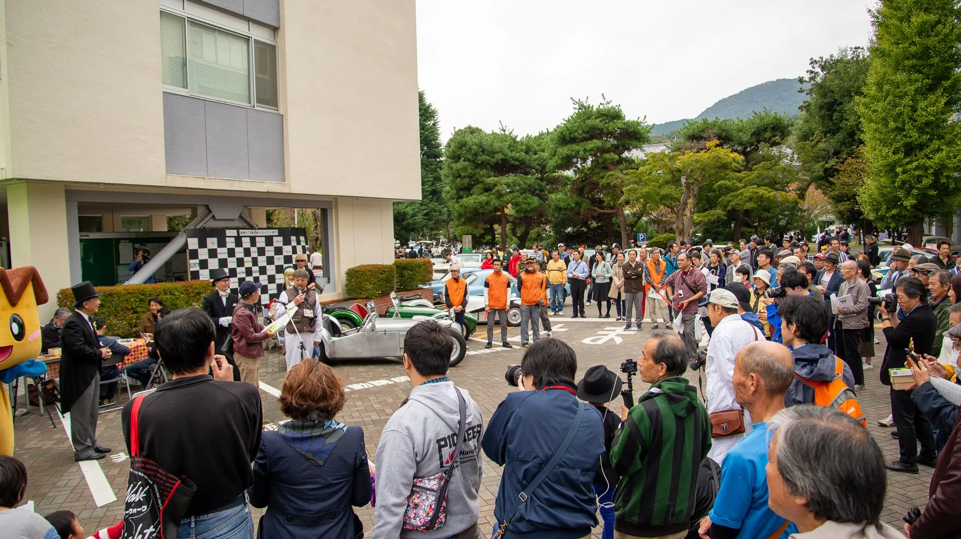 イベントで観客が集まっている様子。クラシックカーが展示され、マスコットキャラクターもいます。