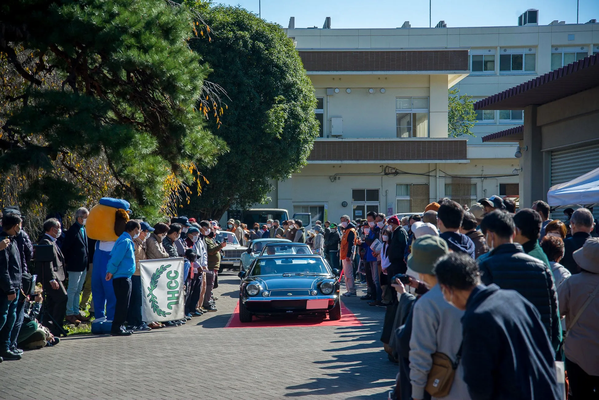 クラシックカーのイベントの様子。多くの観客が見守る中、青いクラシックカーが赤いカーペットの上を走行しています。NHCCと書かれた旗が見えます。