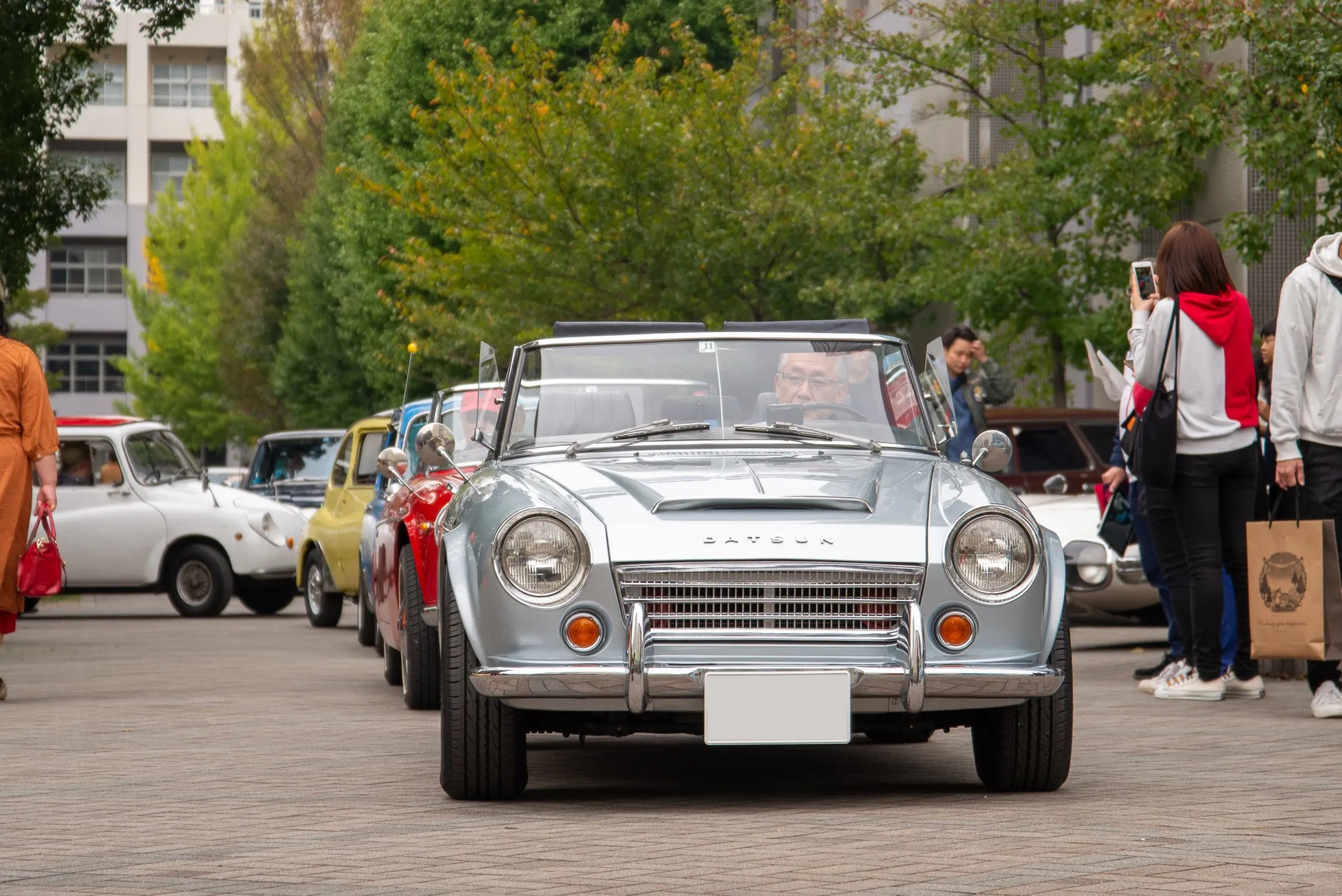 クラシックカーのイベントで、銀色のダットサンが他の古い車と並んで駐車されている様子。観客が車を撮影したり、イベントを楽しんでいる。