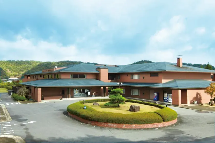 建物の外観写真です。建物の前には、芝生と植え込み、庭木が配置されています。建物の周りには、駐車場と道路があります。空は青く、雲が広がっています。