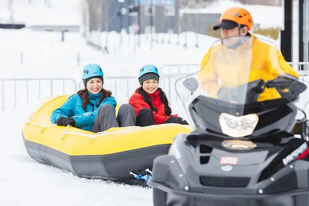 雪の中で、2人の女性が黄色いゴムボートに乗り、スノーモービルに牽引されている様子