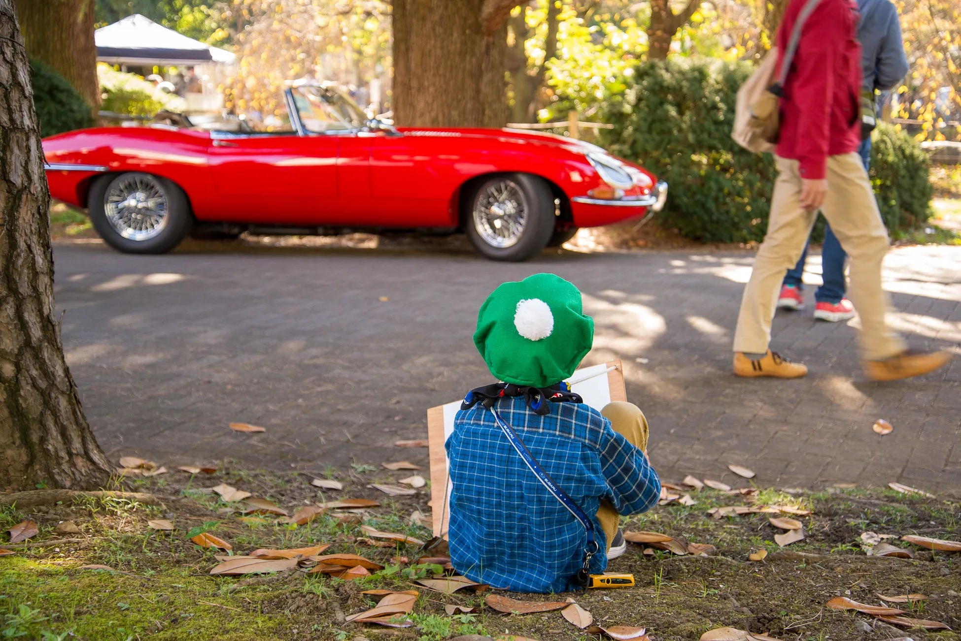 赤いスポーツカーを描くために座っている子供の姿。子供は緑の帽子と青いチェックのシャツを着ています。背景には、歩いている人々、木、そして日陰のテントがあります。