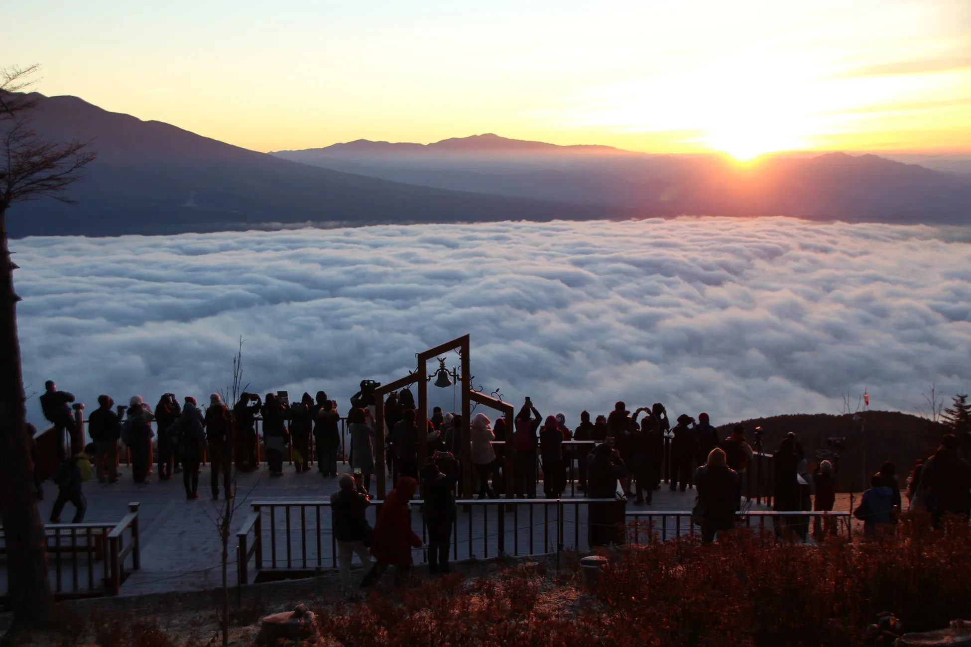 雲海の上に昇る朝日を眺める人々の写真。人々は展望台のような場所に集まり、カメラやスマートフォンでその光景を撮影している。遠くには山々がシルエットとして見え、空はオレンジ色に染まっている。