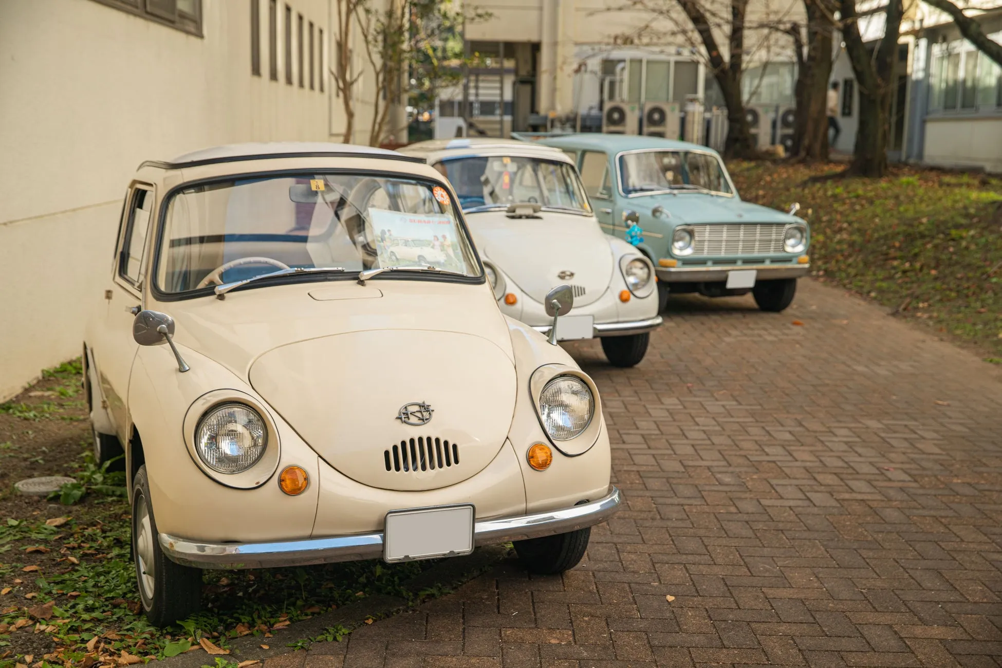 3台のクラシックカーが並んで駐車されている写真。手前にはベージュ色のスバル360、中央には白いスバル360、奥には水色のクラシックカーが写っている。レンガ敷きの道路に車が並び、背景には建物と木々が見える。
