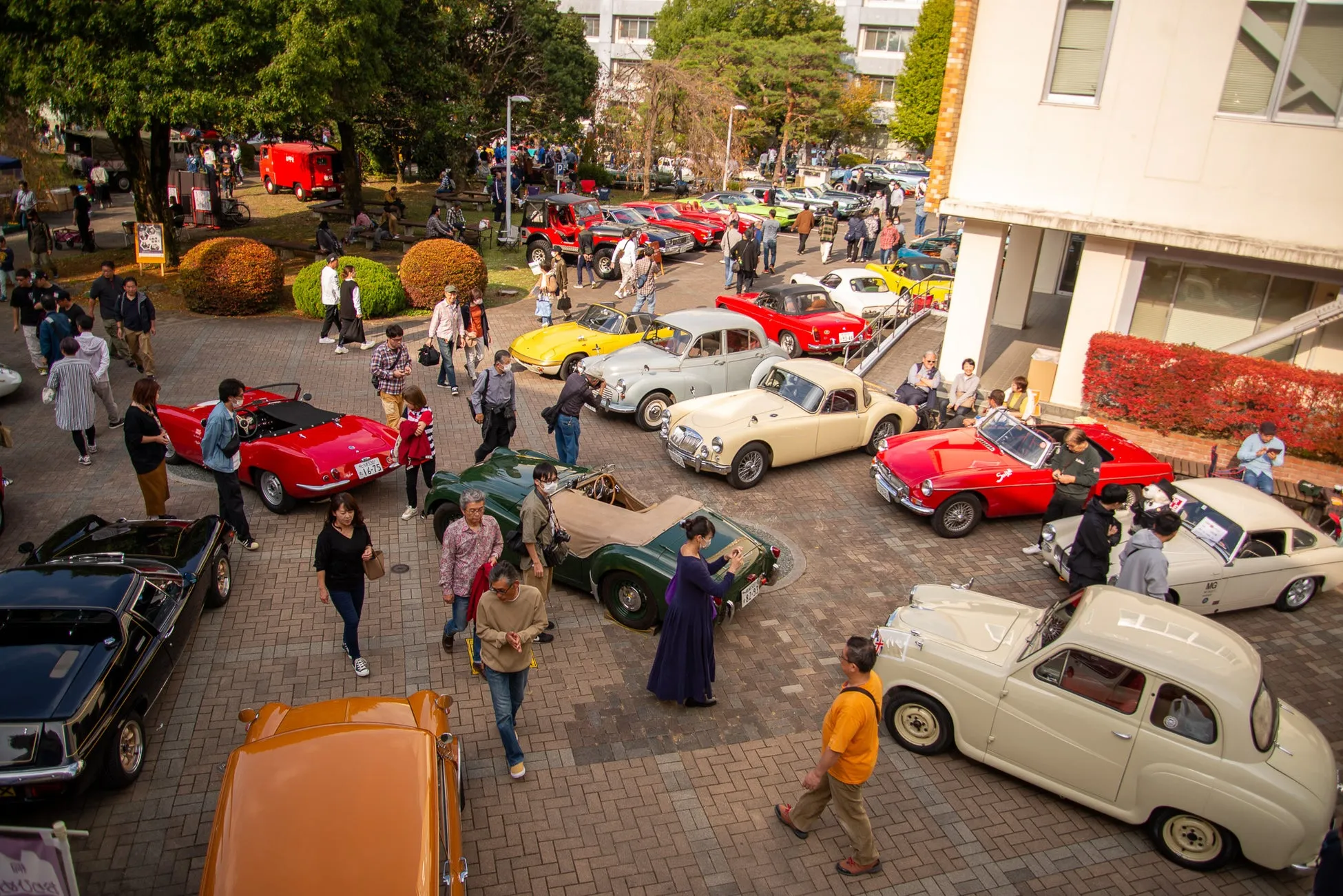 クラシックカーのイベントの様子を捉えた写真。様々な色の古い車が並び、人々がそれらを見たり、写真を撮ったりしている。イベントは屋外で行われ、晴れた日差しの中で活気にあふれている。