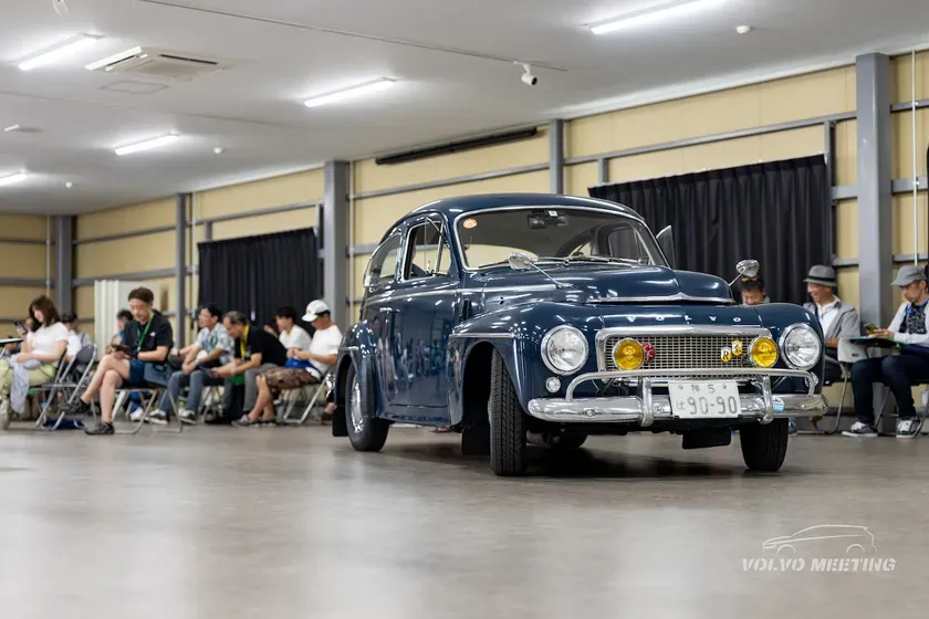 会議室に展示されたクラシックな紺色のボルボ車。周囲には観客が座っており、イベントの様子が伺える。