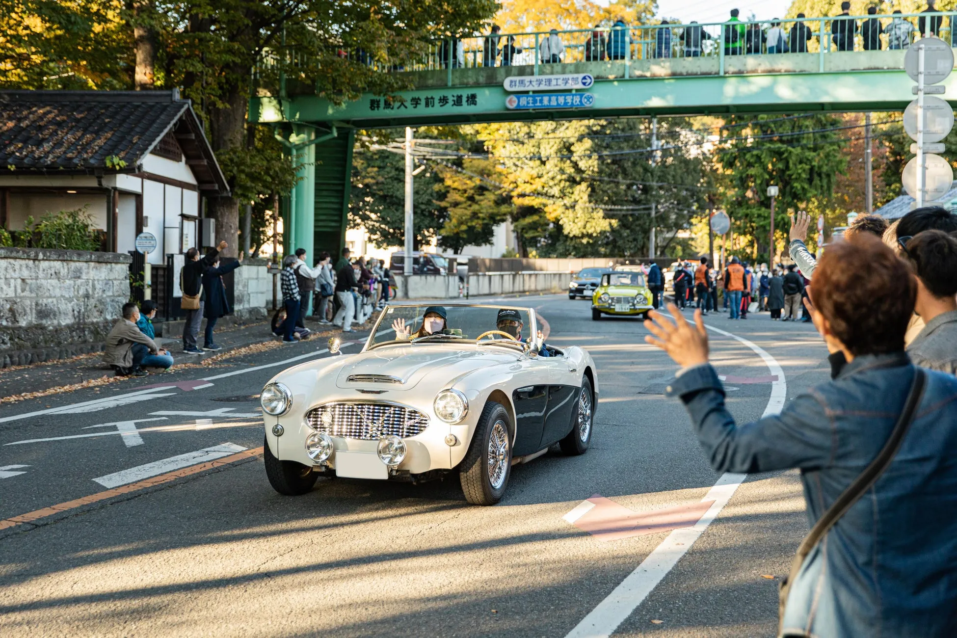 クラシックカーに乗った人々が手を振っており、観客も手を振って応えている様子が写っています。橋には「群馬大学前歩道橋」と書かれています。