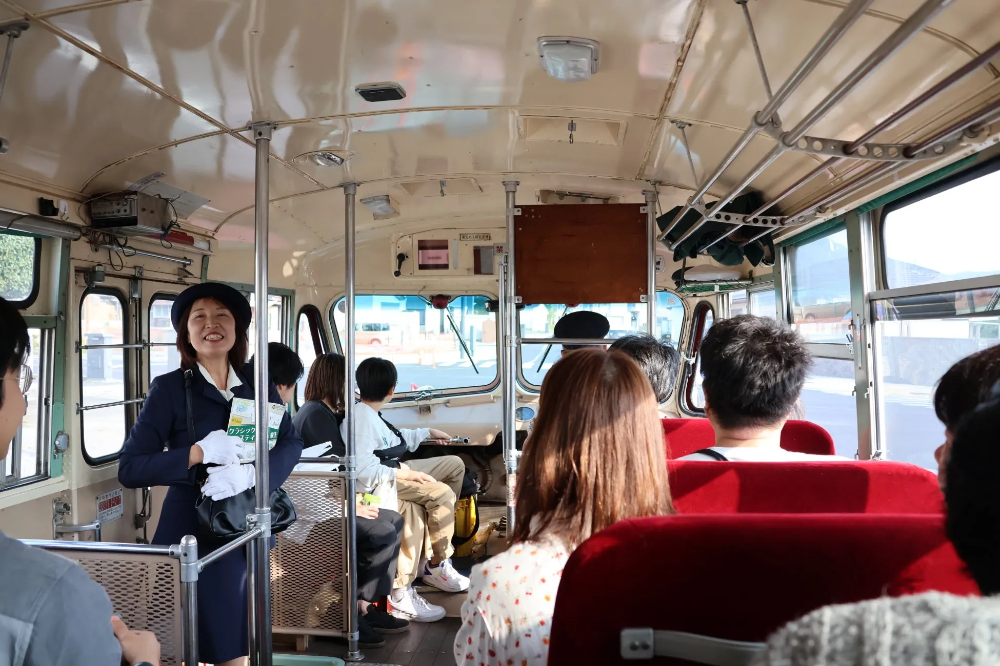 レトロなバスの車内で、制服を着た女性が笑顔で立っており、乗客たちを案内している様子。