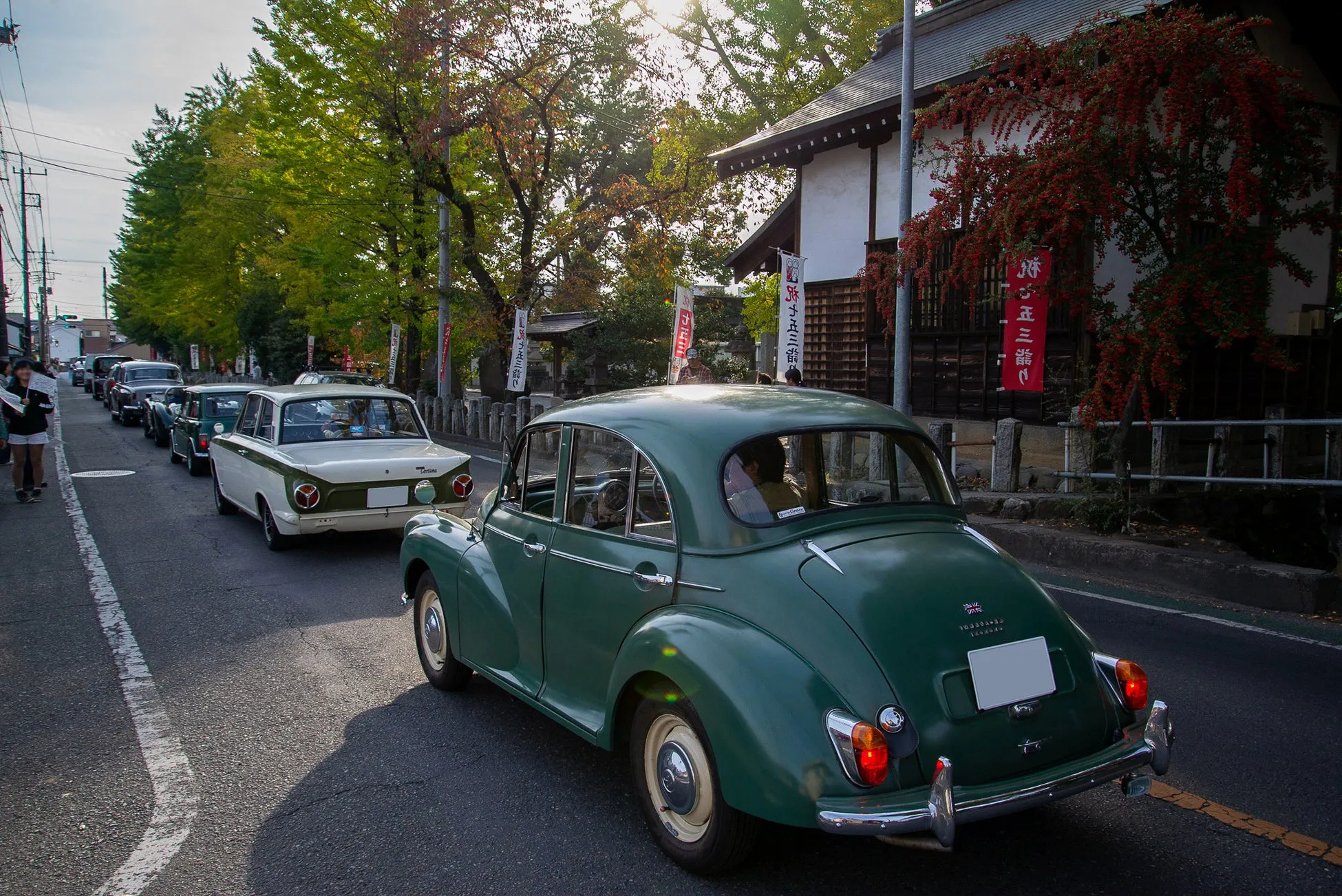 クラシックカーが並んで道路を走行している写真です。緑色のクラシックカーが手前を走行しており、その後ろには他のクラシックカーが連なっています。背景には日本の伝統的な建物と紅葉した木々が見えます。写真全体にノスタルジックな雰囲気があり、イベントやパレードの様子を捉えているようです。