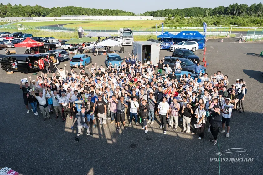 多くの人が集まり、手を振っている写真。背景には車、テント、レーストラックが見える。