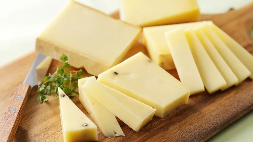 木製のまな板の上に、様々な形にカットされたチーズがいくつかあります。チーズは黄色で、いくつかのチーズには黒い粒が見えます。チーズの近くには、ハーブとチーズナイフがあります。