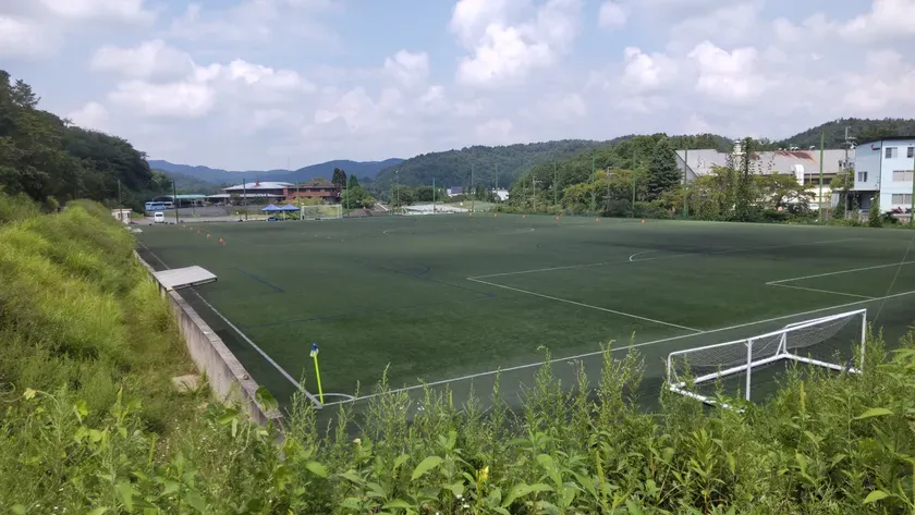 青空の下、サッカー場を撮影した写真。緑の人工芝のフィールドがあり、ゴールポストが設置されている。周囲には木々や建物が見える。