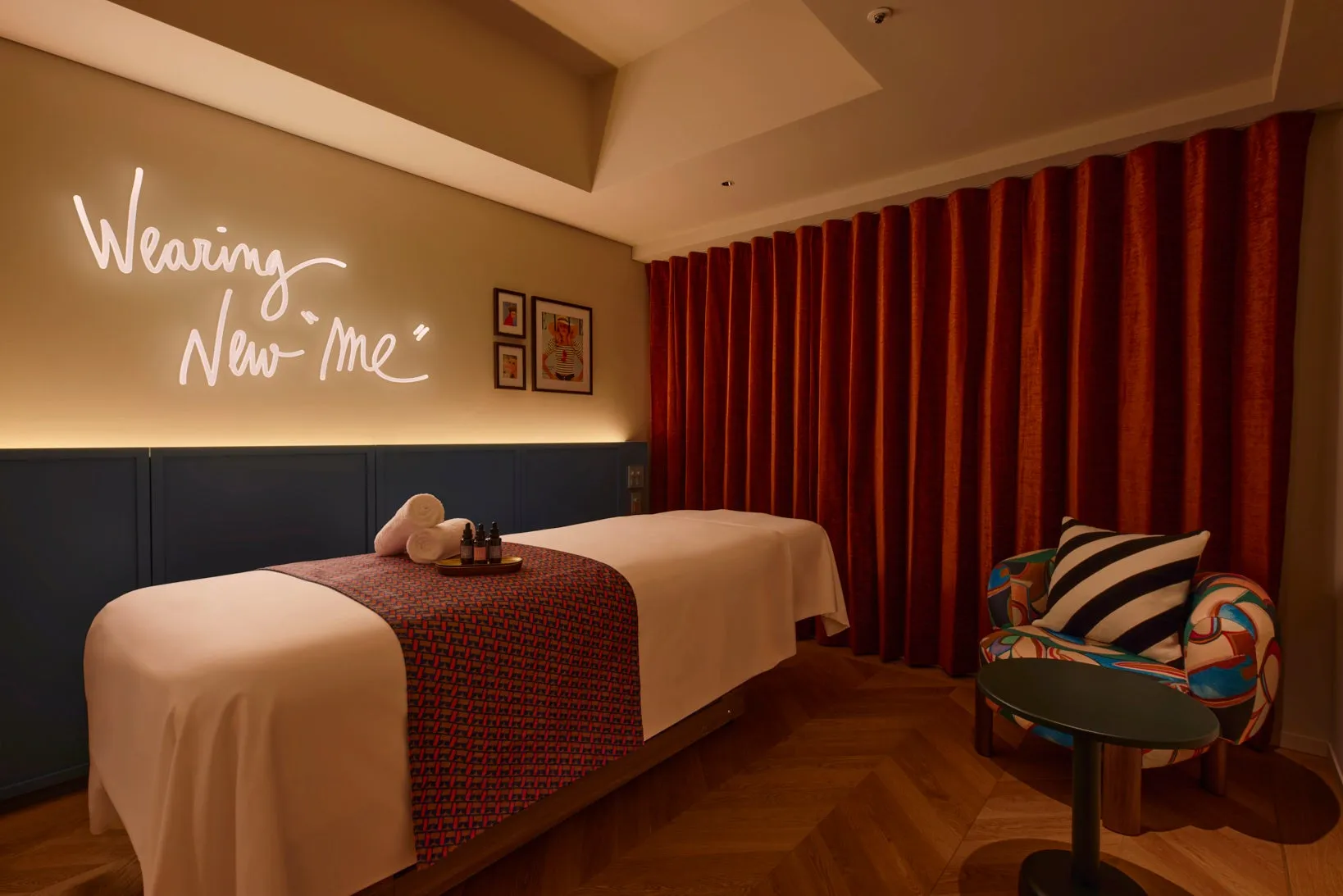 スパトリートメントルーム。ベッド、タオル、美容製品が置かれ、赤いカーテンと椅子がある。壁には「Wearing New me」というネオンサインが飾られている。