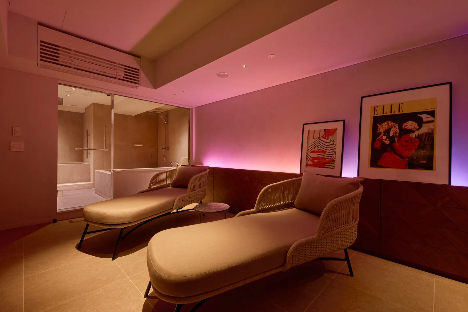 リラックスできるスパルームの写真です。2つのラウンジチェアがあり、ピンク色の照明が施されています。奥にはバスタブとシャワーがあり、雑誌のポスターが壁に飾られています。