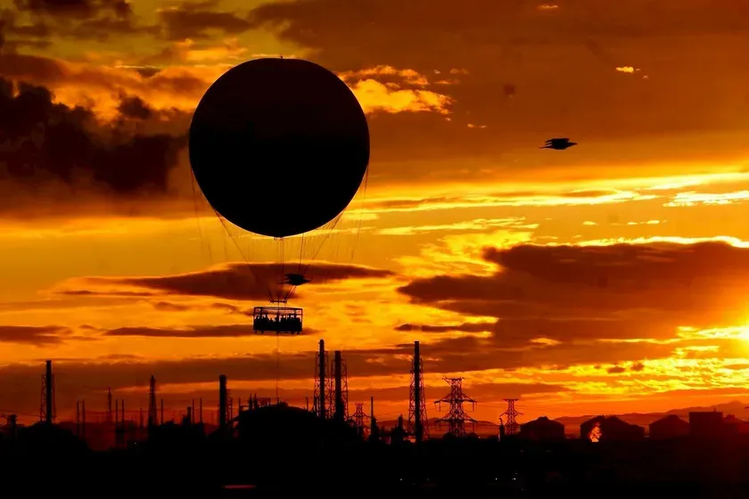 夕焼け空をバックに、熱気球が飛んでいる写真です。熱気球の下にはゴンドラがあり、人が乗っているのが確認できます。前景には工場のシルエットがあり、夕焼けのオレンジ色と対比されています。