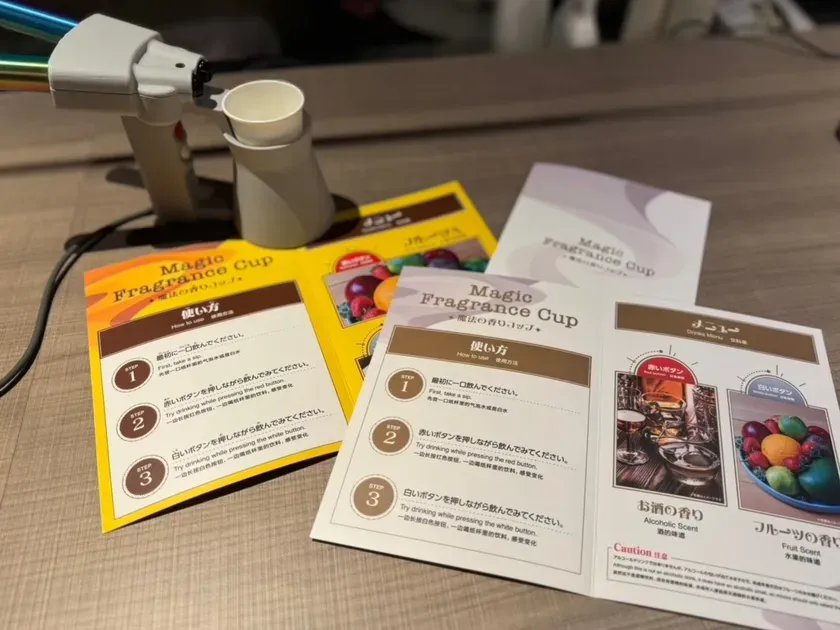 Magic Fragrance Cupのパンフレットと、それを使用するためのデバイスが写っている写真。デバイスは、カップに香りを吹きかけるように見える。