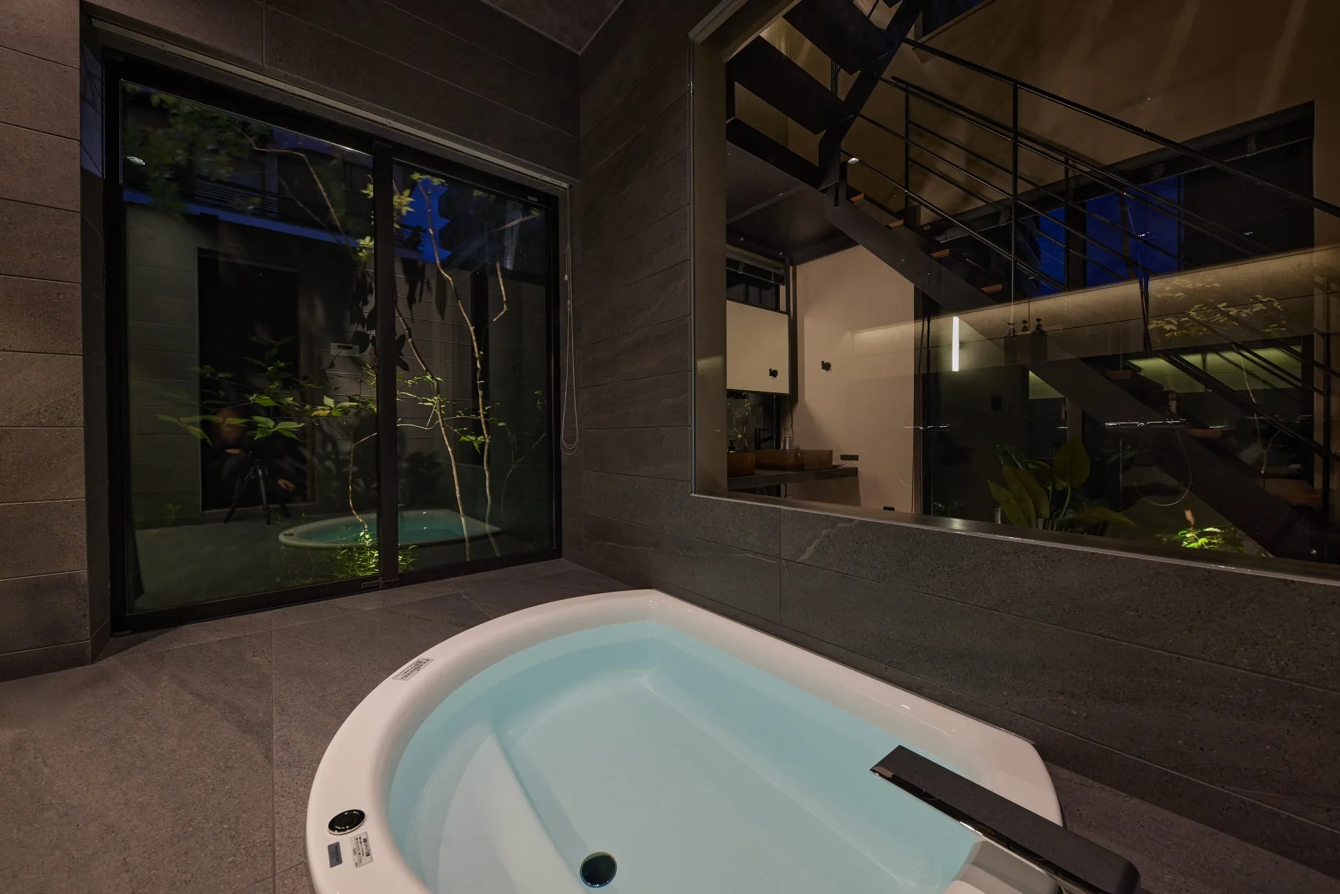 モダンな浴室に白いバスタブがあり、大きな窓からは夜の庭園と小さなプールが見えます。右側の鏡にはスタイリッシュな階段と洗面台が映り込み、高級感とリラックス感あふれる空間を演出しています。