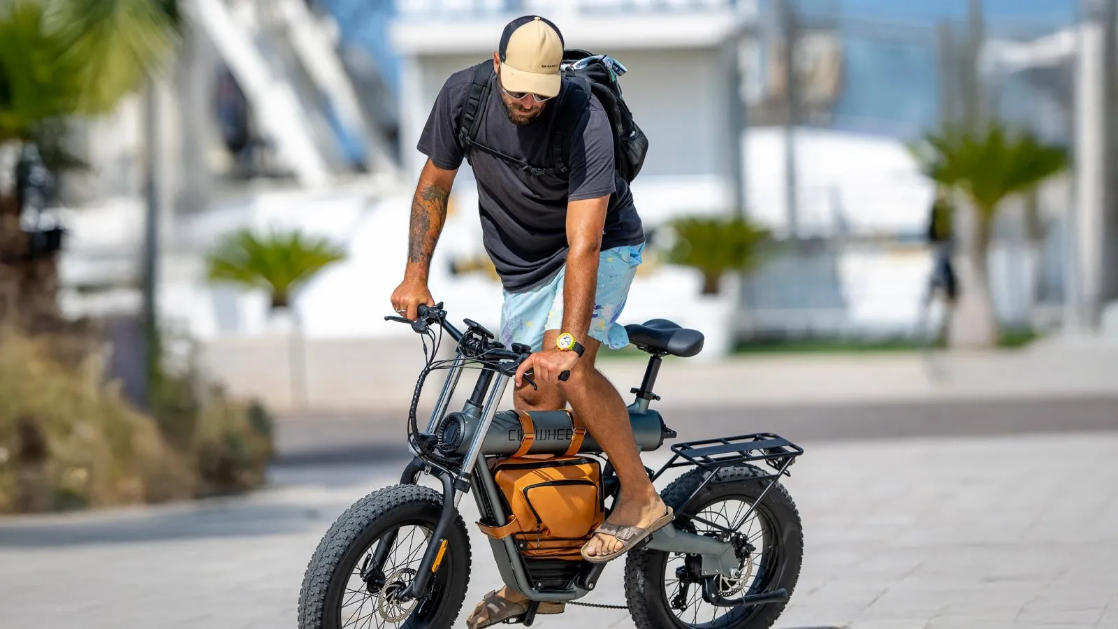 男性が電動自転車に乗っている写真。男性はサングラス、帽子をかぶり、リュックサックを背負っている。電動自転車は茶色のバッグが付いている。