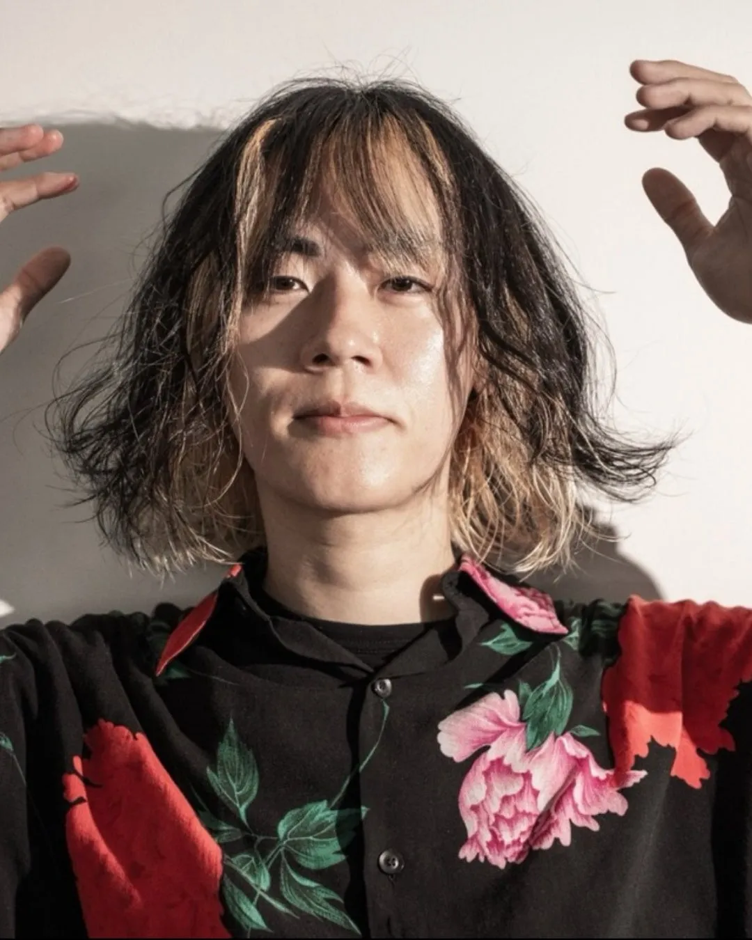 男性がカメラ目線で、両手を頭の横に上げています。彼は黒いシャツを着ていて、赤い花柄が特徴です。彼の髪は黒と金髪のツートンで、顔は少し汗ばんでいます。