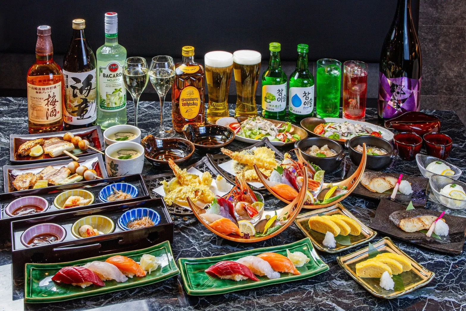 テーブルいっぱいに料理が並べられた写真。寿司、刺身、揚げ物、焼き鳥など、様々な日本食が写っています。飲み物も豊富で、ビール、焼酎、カクテルなどがあります。