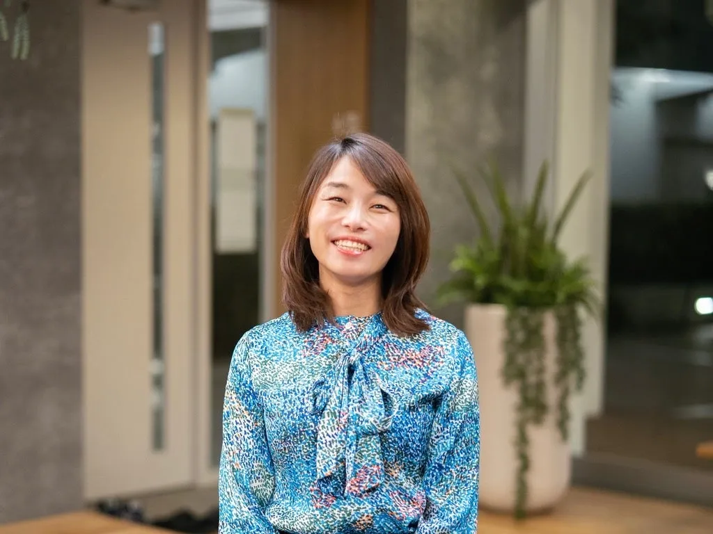 笑顔の女性のポートレート。彼女は青い柄のブラウスを着ており、カメラに向かって微笑んでいます。背景には緑の植物とモダンなインテリアが見えます。