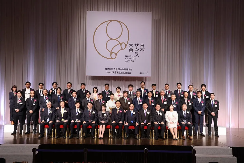 表彰式で記念撮影をする人々の写真。背景には「NIHON SERVICE AWARD」のロゴと日付が表示されている。