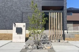 家の玄関前にある植栽の様子を写した写真です。庭には、木と石が配置され、モダンな外観の家を引き立てています。