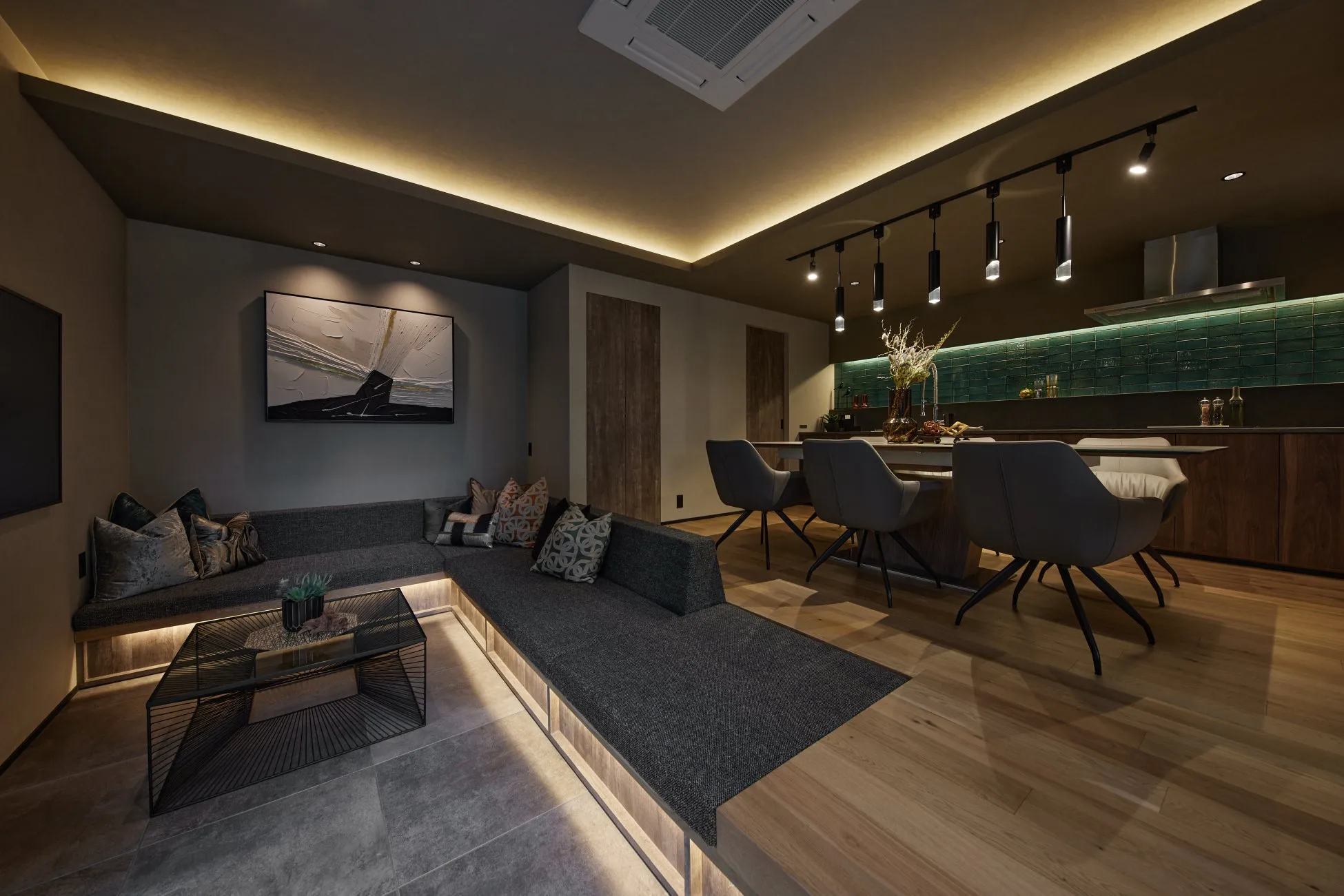 L字型ソファのあるリビングエリアと、ダイニングテーブル、オープンキッチンが一体となったモダンでスタイリッシュな空間です。間接照明が落ち着いた雰囲気を演出し、壁のアートやデザイン性の高い家具が洗練された印象を与えています。