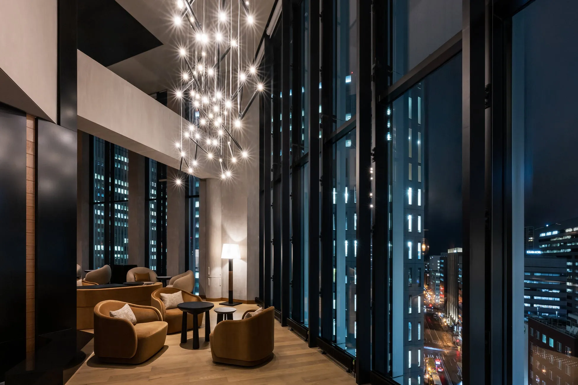 夜の街並みを望むモダンなラウンジのインテリア写真。シャンデリアが輝き、窓からは高層ビル群の明かりが映し出されている。茶色のソファと丸テーブルが配置され、落ち着いた雰囲気を醸し出している。