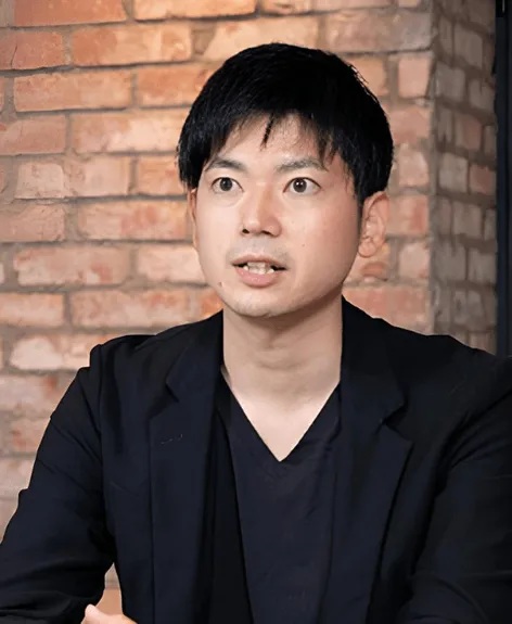 レンガの壁を背景に、ジャケットを着たアジア系の男性のポートレート。彼はカメラを見て、話しているように見える。