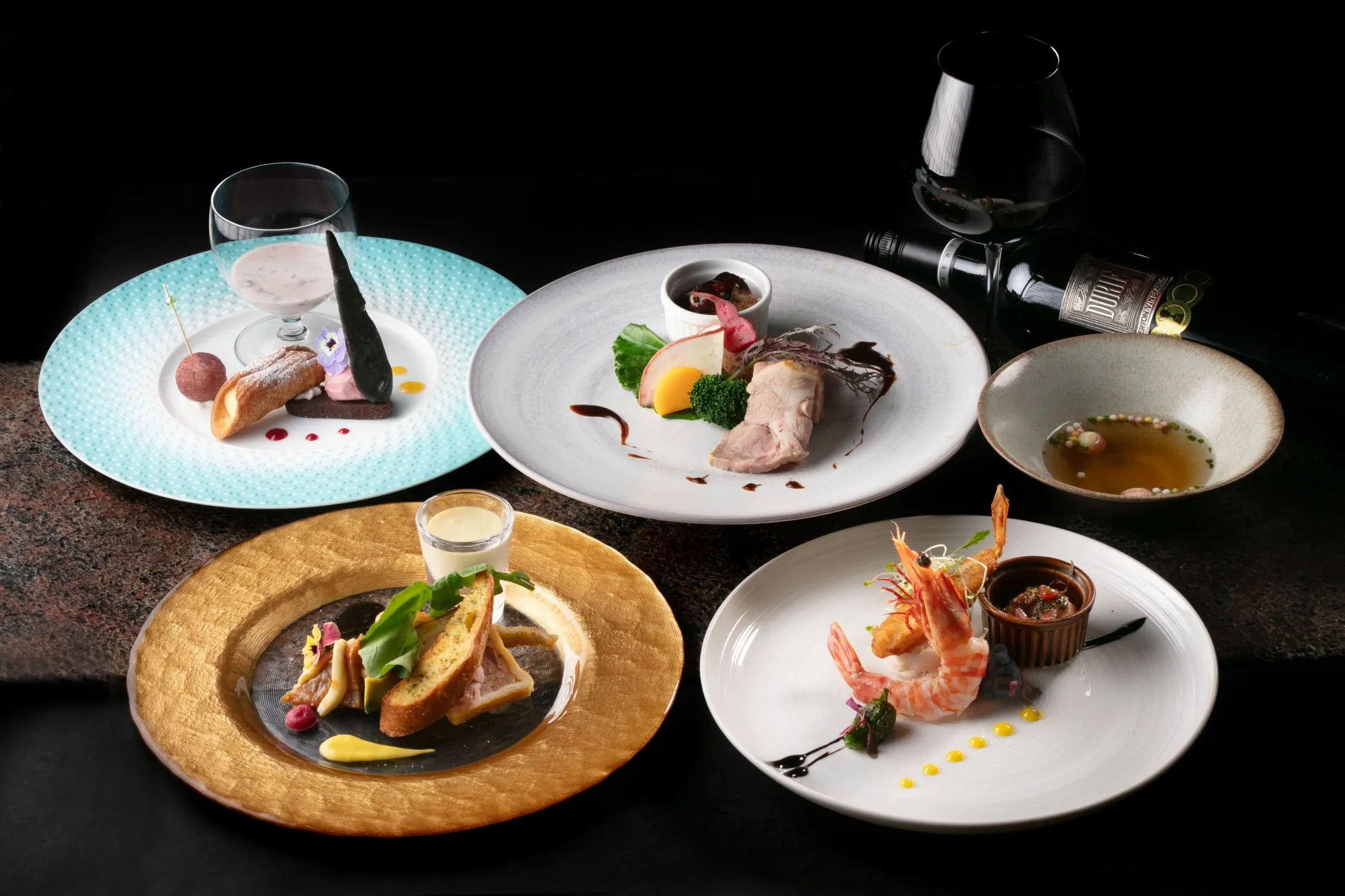 様々な料理が盛り付けられたコース料理の画像。デザート、スープ、前菜、メイン料理、エビ料理など、様々な料理が美しく配置されており、ワインボトルとワイングラスも添えられている。