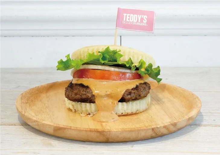 木製の皿に盛られた、具材が豊富なハンバーガーです。ジューシーなパティにはとろりとしたソースがかかり、新鮮なレタス、トマト、オニオンが挟まれています。バンズには「TEDDY'S BIGGER BURGERS™」と書かれたピンクの旗が刺さっています。
