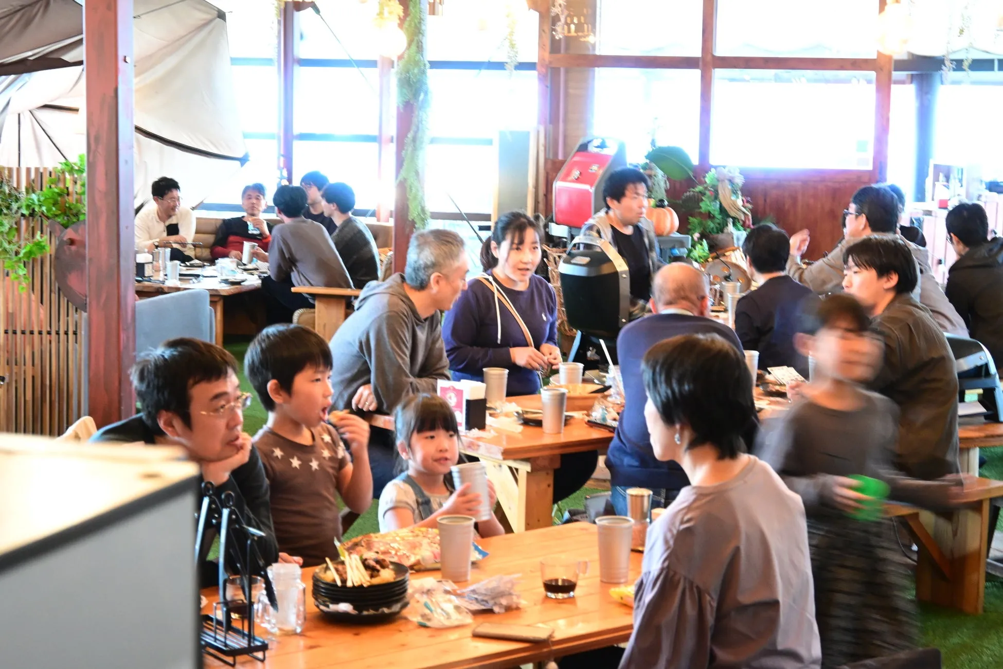 レストランで食事を楽しむ人々の様子を捉えた写真。木製のテーブルと椅子が配置され、人々は食事をしたり、会話を楽しんだりしている。背景には窓があり、自然光が差し込んでいる。