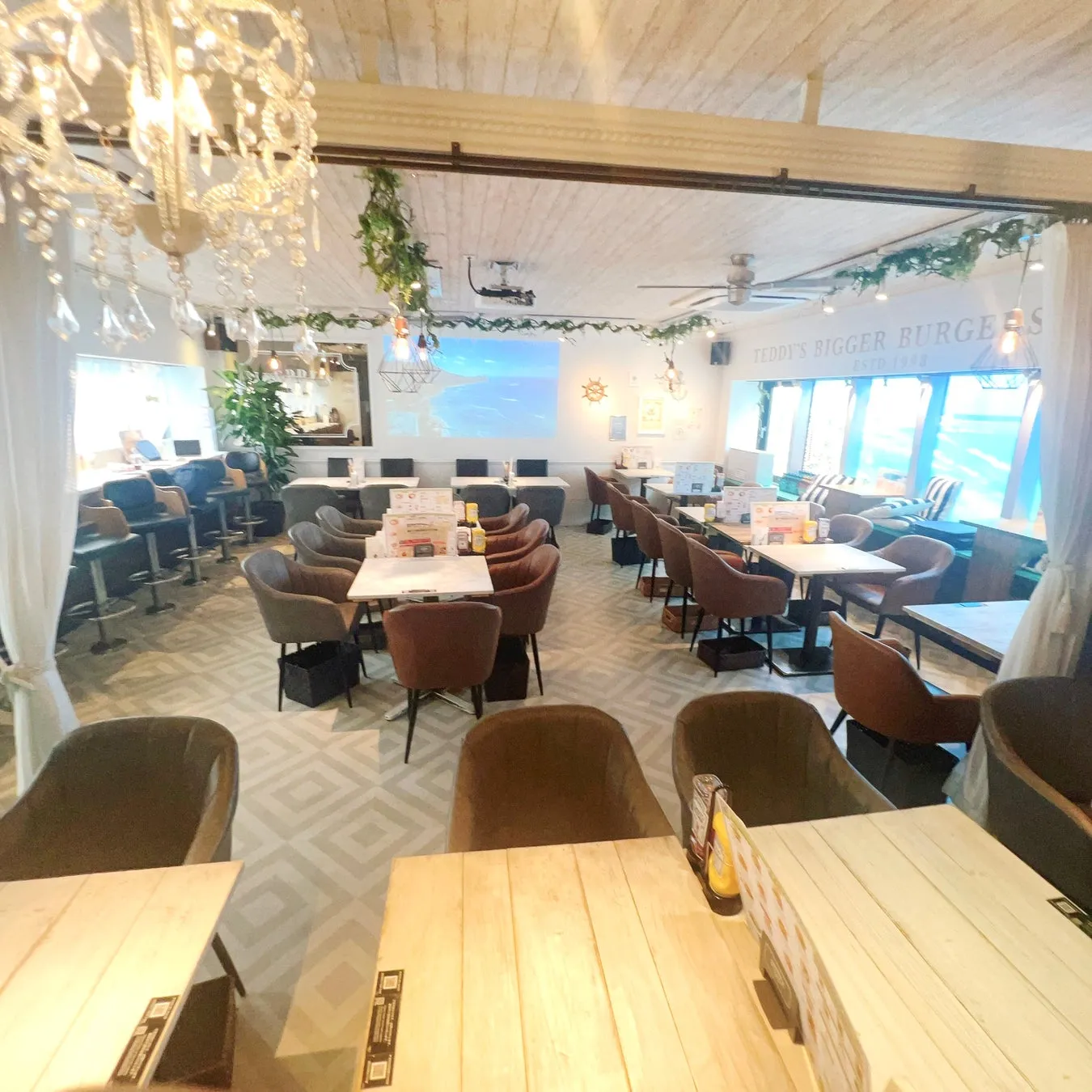 広々としたレストランの店内。ライトウッドのテーブルと落ち着いた色の椅子が多数配置され、モダンでカジュアルな雰囲気が漂う。壁にはビーチの映像が投影されており、ハワイアンな要素も感じられる空間。