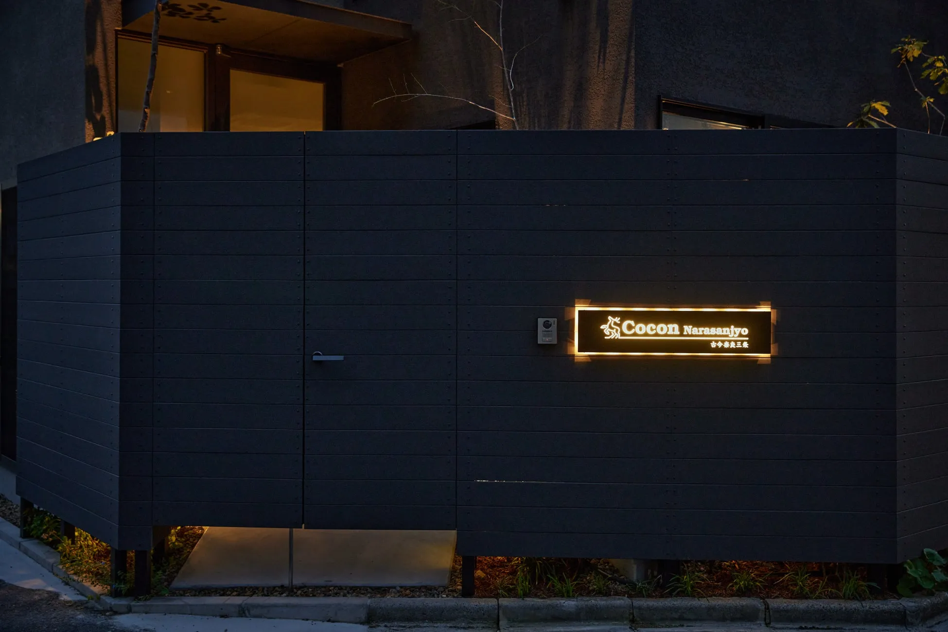 夜のモダンな建物の入り口を捉えた画像です。黒い壁に光る「Cocon Narasanjyo 古今奈良三条」の看板が設置されており、足元には間接照明が施されています。