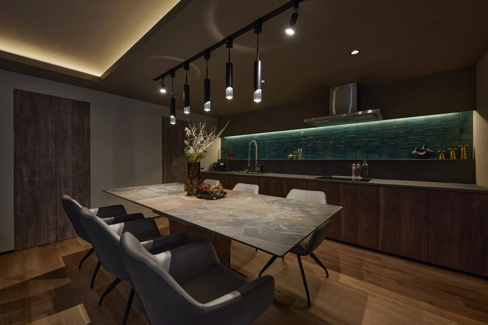 モダンで洗練された雰囲気のダイニングキッチン。大理石調の大きなテーブルとスタイリッシュな椅子が配置され、天井のトラックライトと間接照明が空間を照らしています。グリーンのタイルが印象的なキッチンスペースには、ダークウッドのキャビネットが備え付けられています。