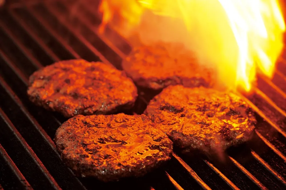 熱いグリルで炎を上げて焼かれている複数のハンバーグが写っています。こんがりと焼き色がつき、肉汁が滲み出ており、食欲をそそる調理中の様子が伝わってきます。