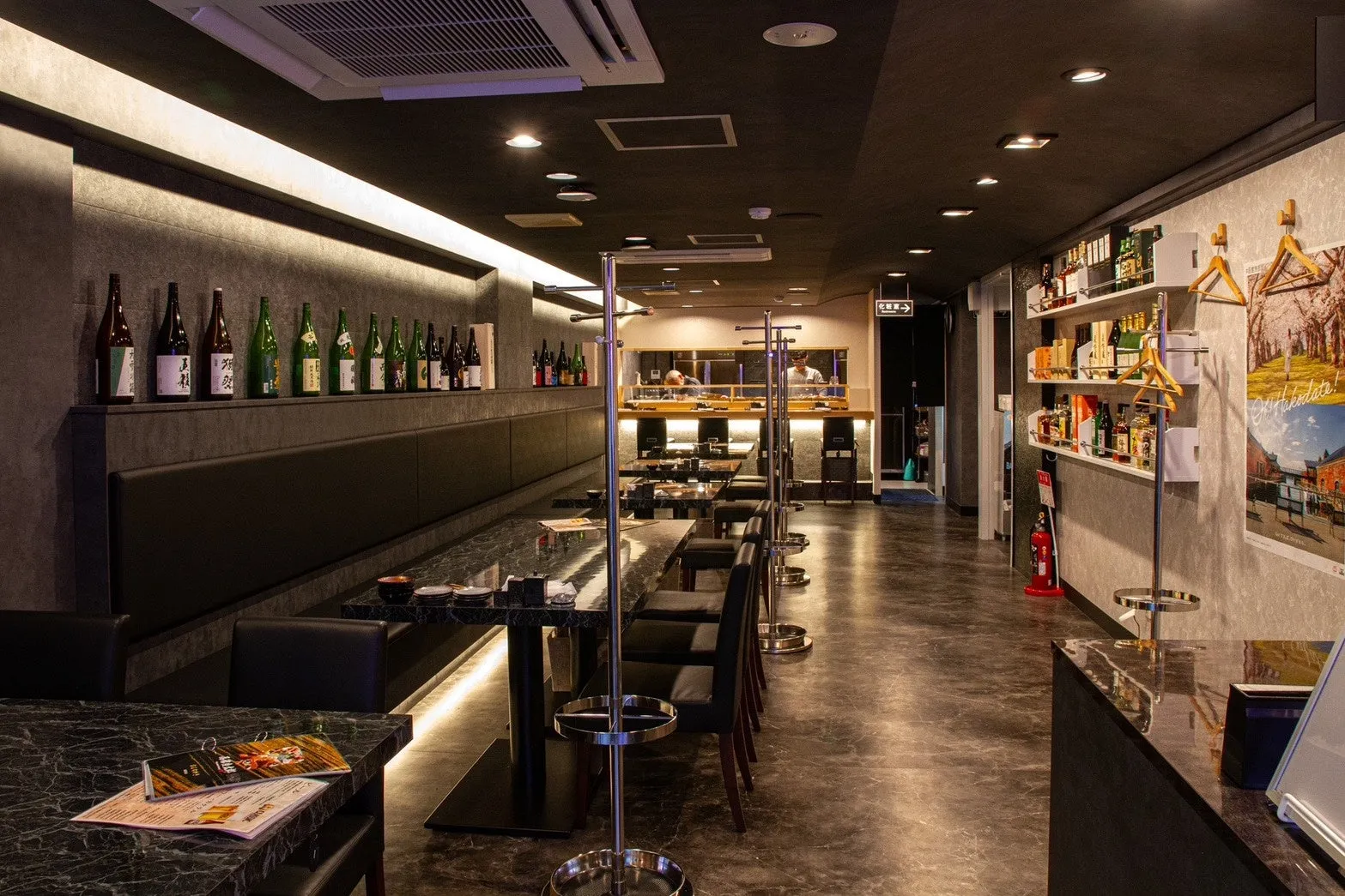 店内を写した写真。テーブル席が並び、奥にはカウンター席と厨房が見える。壁にはお酒の瓶が飾られ、お店の雰囲気を醸し出している。