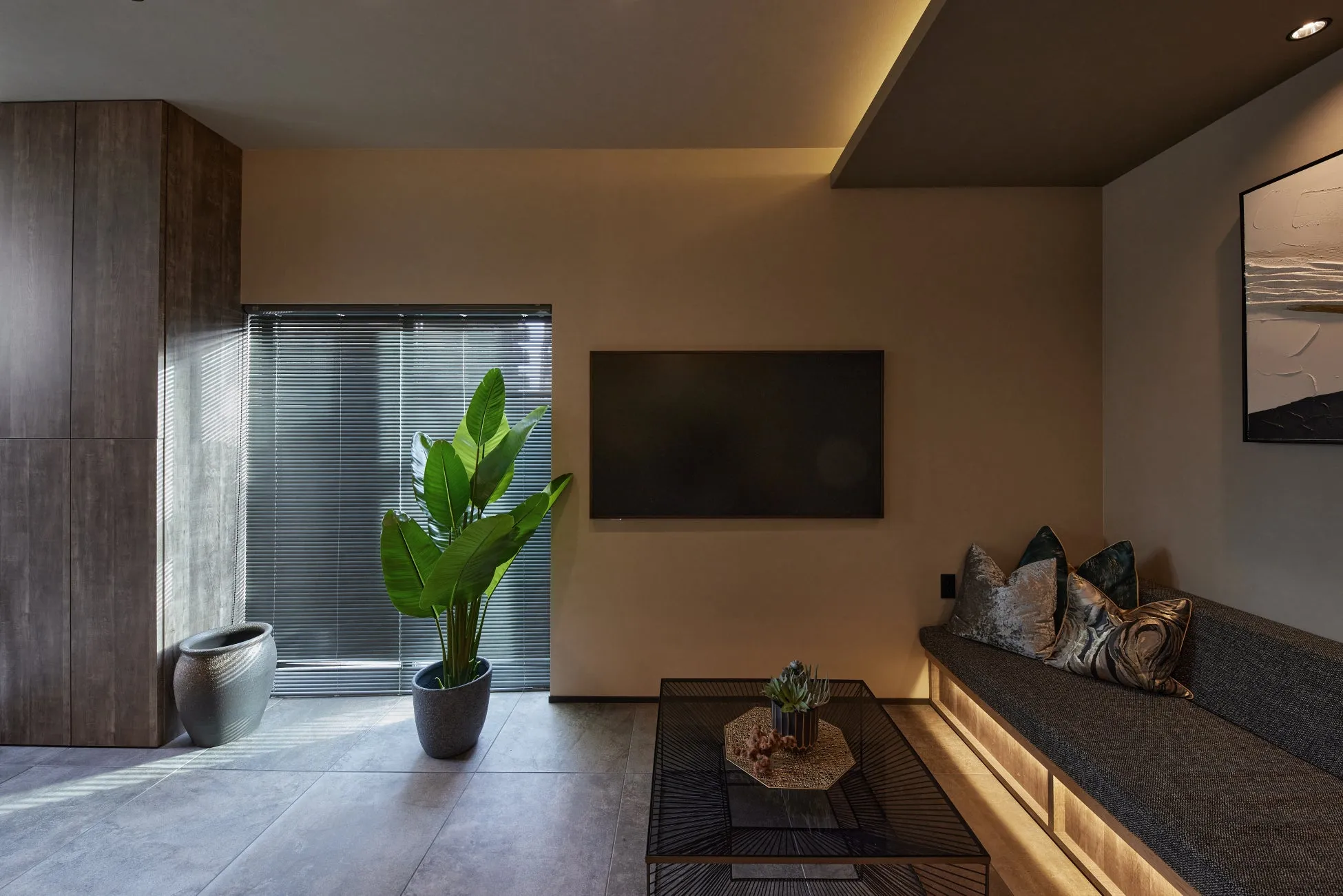 モダンで落ち着いた雰囲気のリビングルーム。壁掛けテレビ、大きな観葉植物、スタイリッシュなソファとコーヒーテーブルが配置され、間接照明が洗練された空間を演出しています。