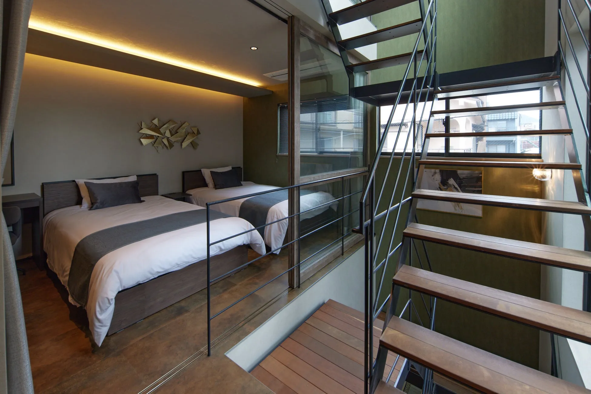 モダンな寝室とスタイリッシュな階段が一体となった空間の画像です。ツインベッドが配置され、間接照明が温かい雰囲気を作り出しています。木と金属を組み合わせた階段が特徴的で、都会的な宿泊施設を思わせる洗練されたインテリアです。