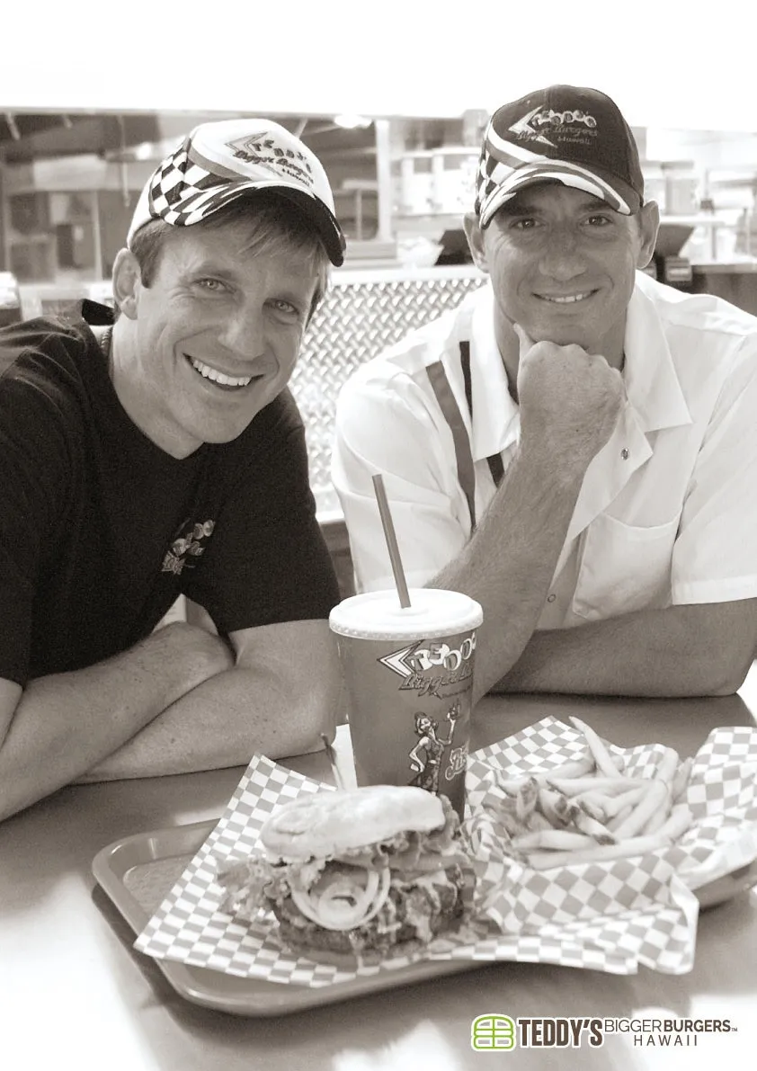 モノクロ写真で、笑顔の男性二人がレストランのテーブルに座っています。テーブルにはハンバーガー、フライドポテト、ドリンクが置かれており、テディーズビガーバーガーズの食事を楽しんでいる様子が伺えます。