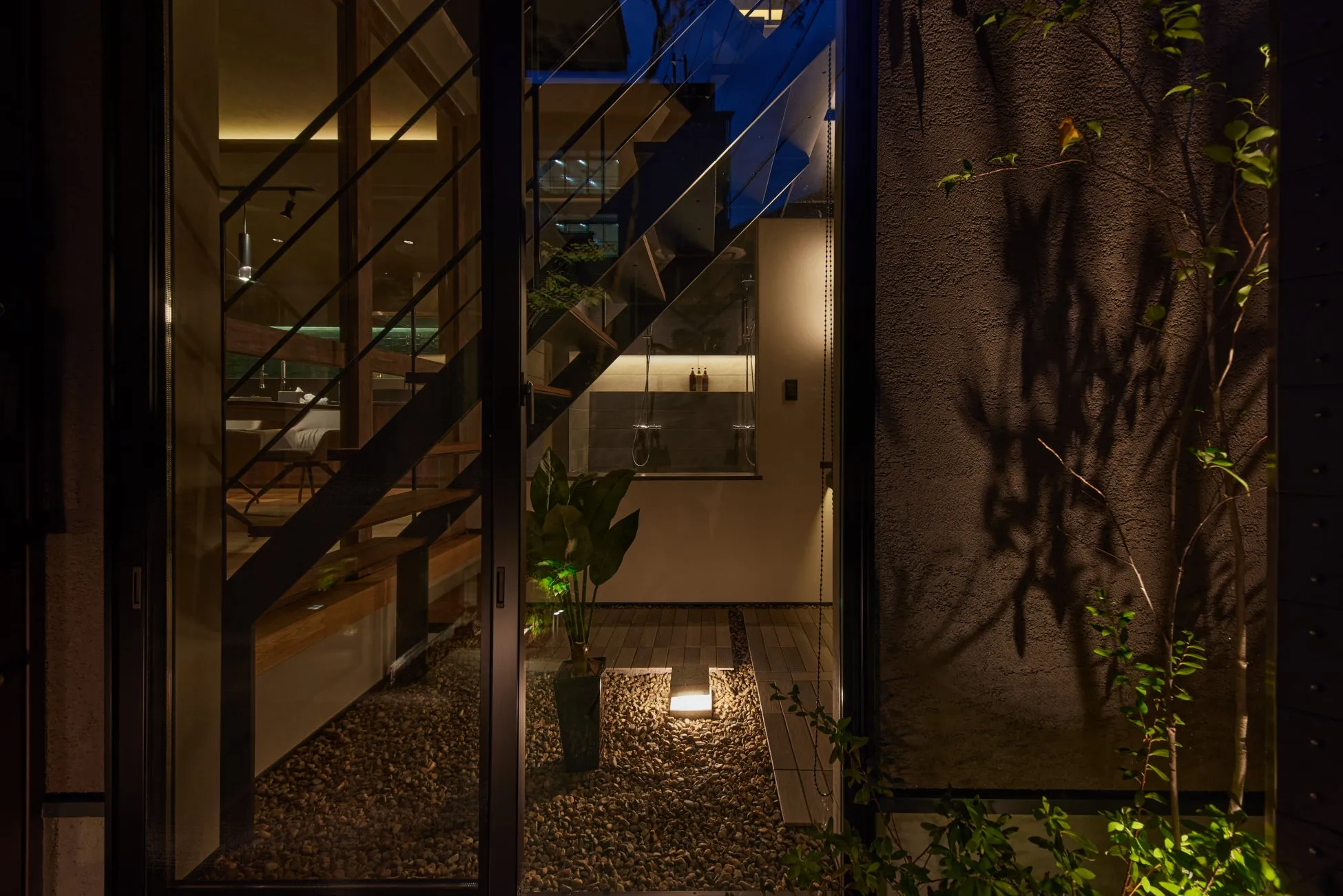 ガラス越しに見える、モダンなデザインの家の外観。階段やシャワー室、植木鉢などが配置され、夜間の照明が美しい。