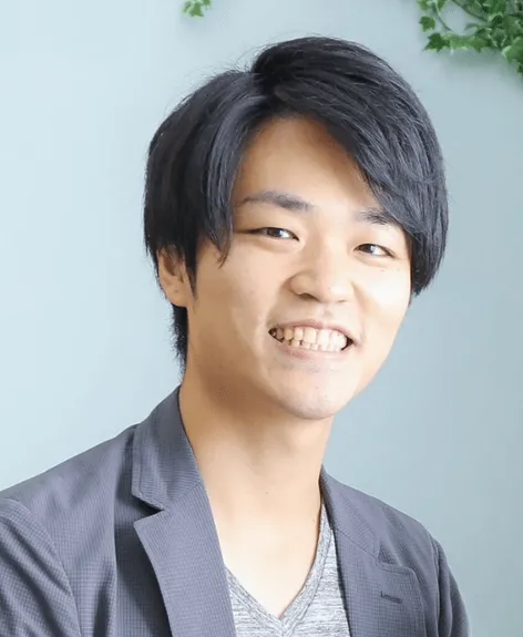 笑顔の男性のポートレートです。彼はジャケットとインナーを着ており、カメラに向かって微笑んでいます。背景は明るい青色で、植物が少し見えます。