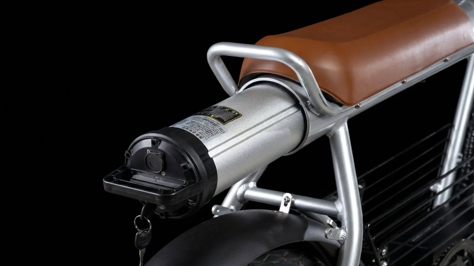 電動バイクのバッテリー部分のクローズアップ写真。バッテリーは銀色で、黒いカバーが付いています。バイクのフレームとシートの一部も見えています。