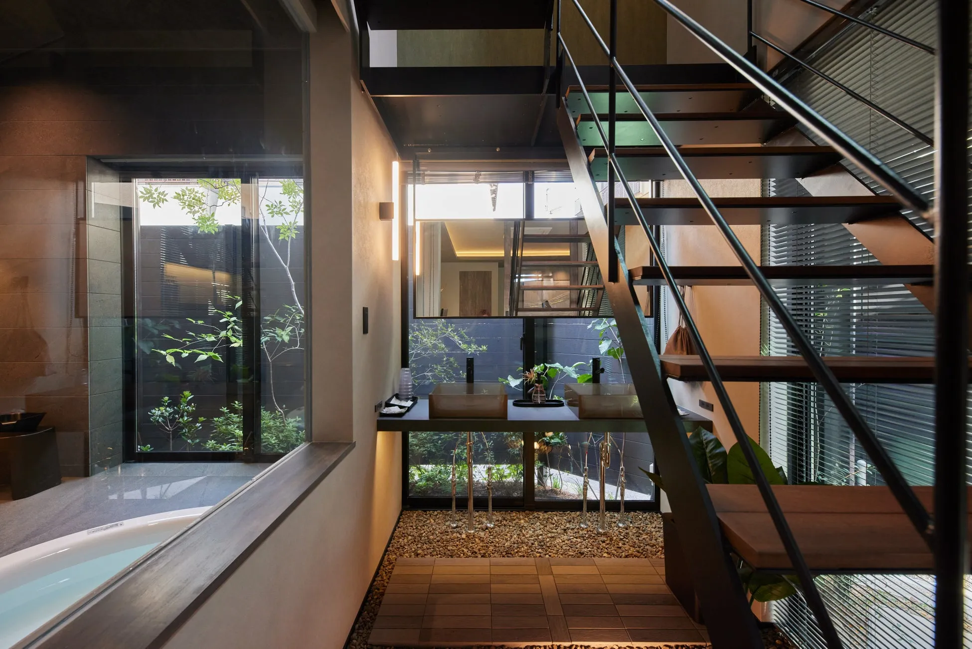モダンで洗練されたデザインの住宅内部。バスタブ、二つの洗面台、黒い金属製の階段が配置され、大きな窓からは豊かな緑が望める開放的な空間です。
