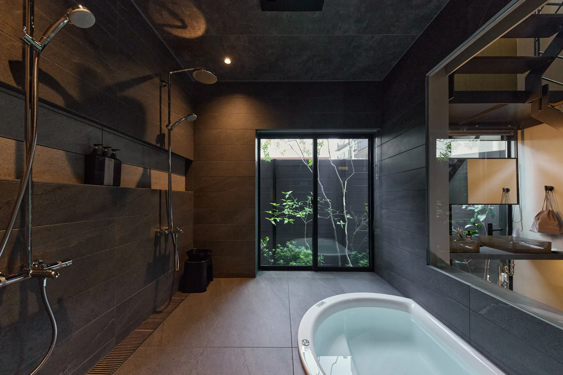 モダンでスタイリッシュな浴室の全景です。深いグレーのタイルが壁と天井を覆い、中央には大きな窓から緑豊かな中庭が見えます。白いバスタブと複数のシャワーがあり、落ち着いた照明がリラックスできる空間を演出しています。
