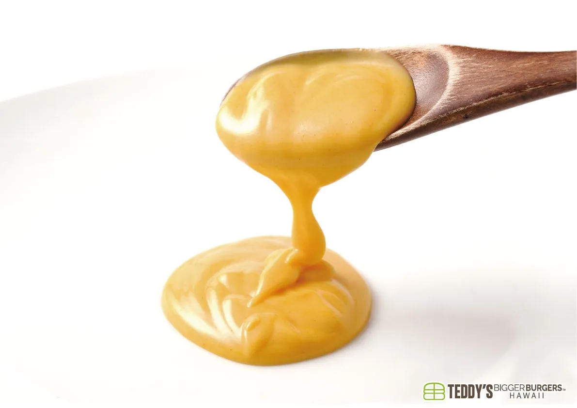 白い背景の上で、木製のスプーンからとろりとした黄色いソースが流れ落ち、下に溜まっている様子を捉えたクローズアップ画像です。ソースは濃厚でクリーミーな質感をしています。