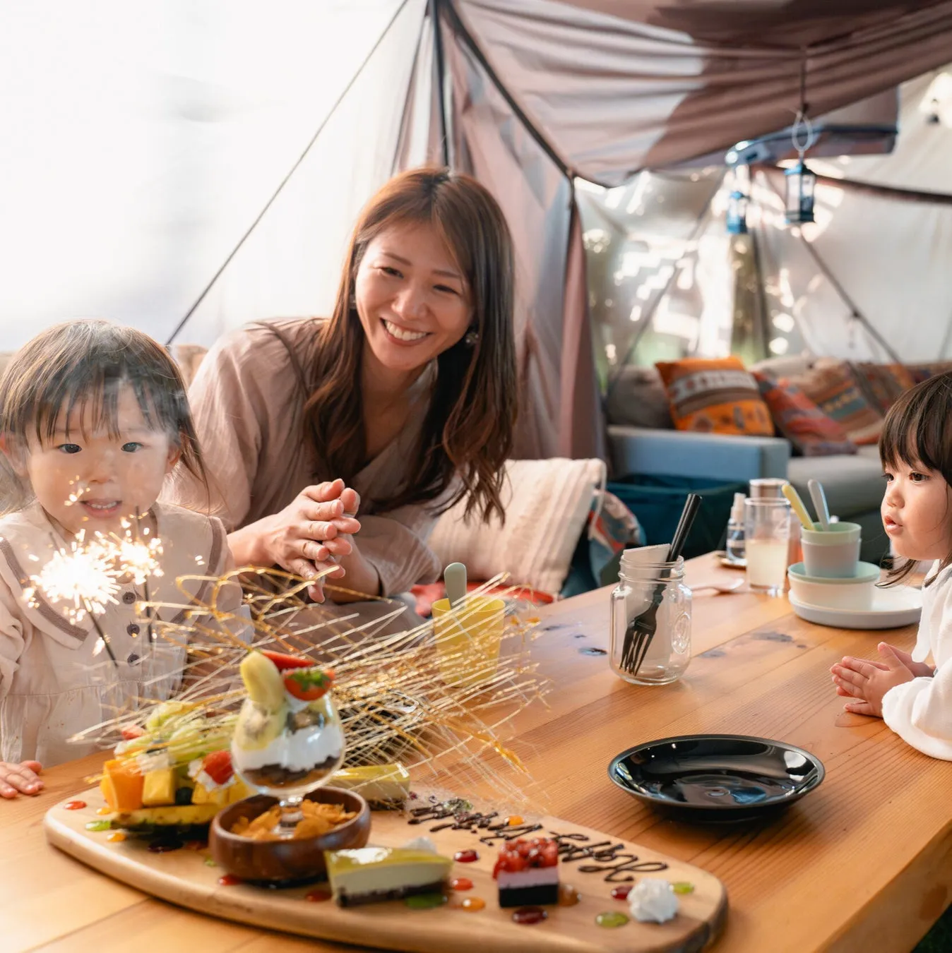 テーブルを囲んで楽しそうにしている家族の写真。子供たちは花火に興味を示し、母親は笑顔でそれを見守る。テーブルには美味しそうな料理が並べられ、特別な日のように見える。