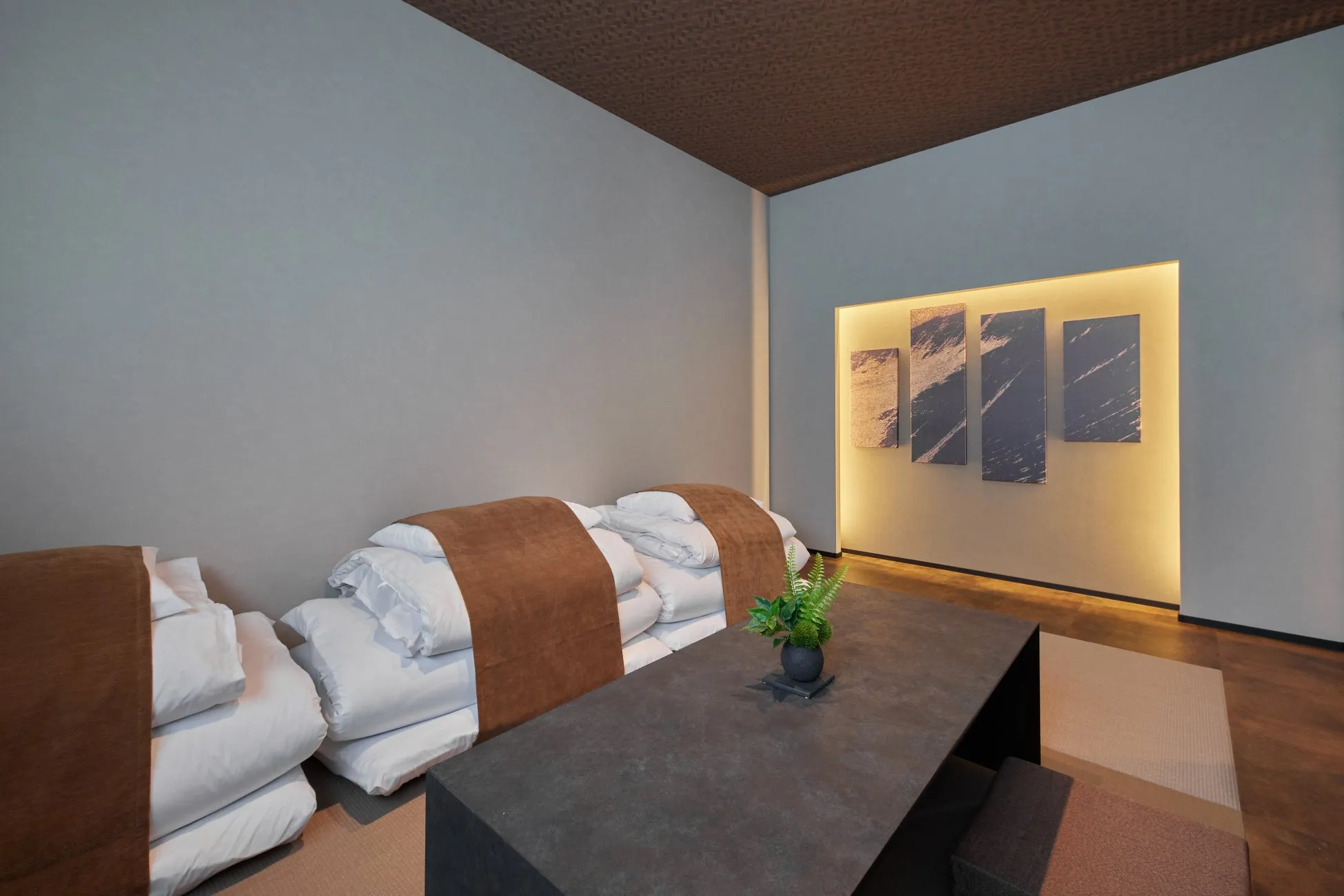 グレーの壁に沿って整然と積み重ねられた布団と、中央に置かれたシンプルでモダンなテーブルがある和モダンの室内です。奥の壁には間接照明で照らされたアートパネルが飾られ、落ち着いた雰囲気を醸し出しています。