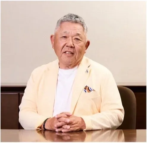 テーブルに座り、カメラ目線で微笑む高齢男性のポートレートです。彼は白のインナーに薄い黄色のジャケットを着ています。